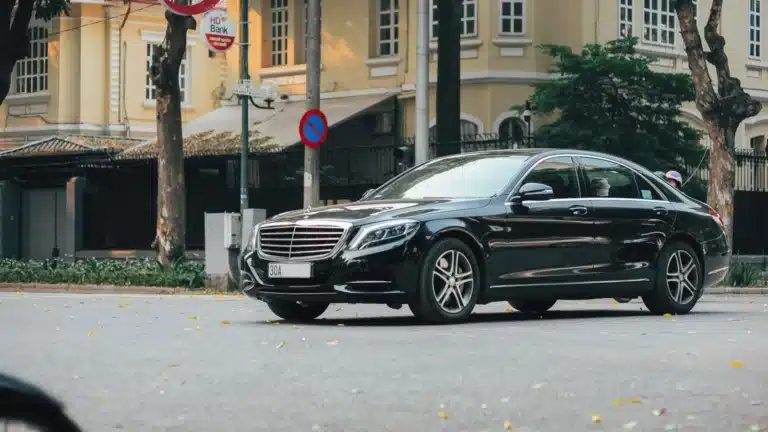 A sleek black sedan navigates an urban street lined with historic buildings and lush trees.