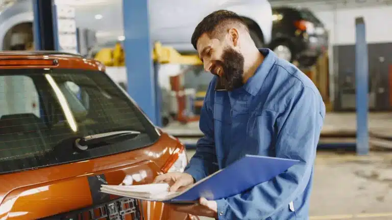 Smiling auto mechanic inspects a car in a service garage, wearing coveralls and holding documents.