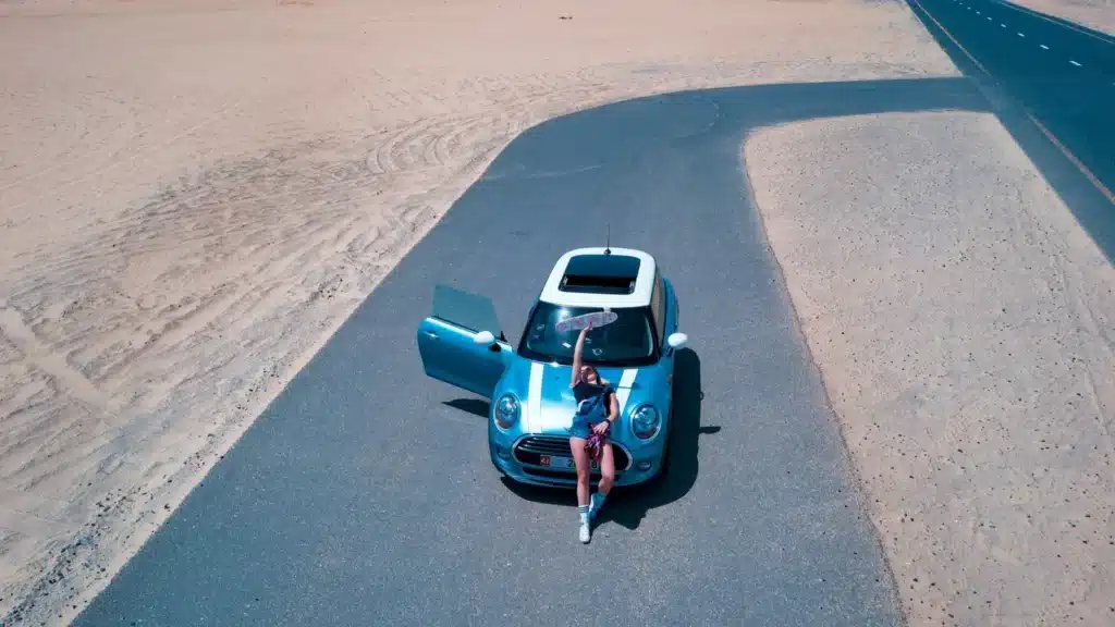 Woman with Mini Cooper on desert road near Dubai. Aerial shot captures vast sandy landscape.