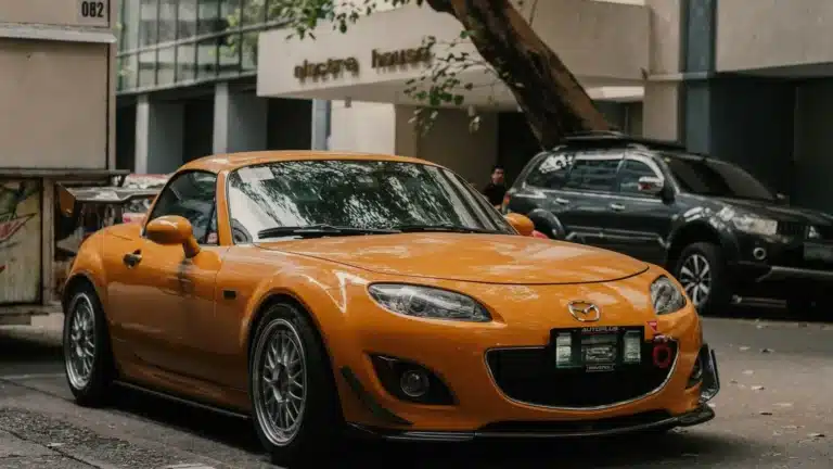 Orange Mazda sports car parked on a city street in front of office buildings, showcasing urban lifestyle.